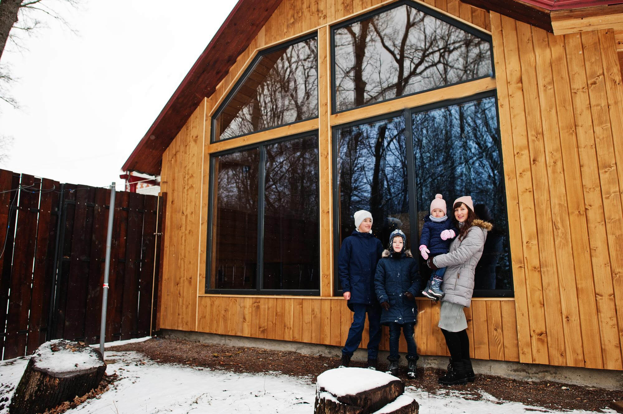 A mother and her 3 children stand in front of a large wooden cabin with a glass window. The family is dressed warmly in winter coats, hats and gloves. There's snow on the ground around them.